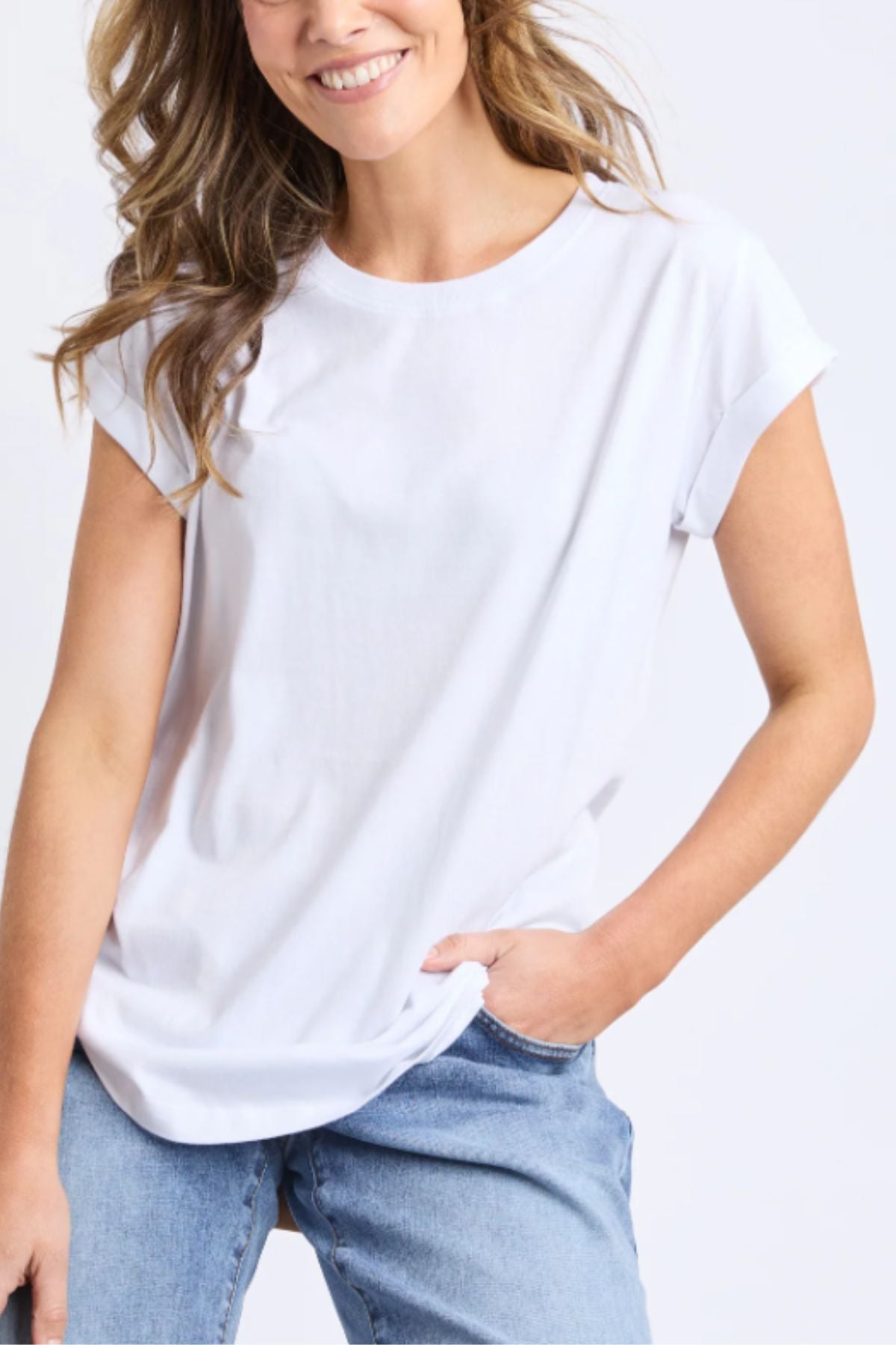 A smiling woman with wavy hair wears the Foxwood Manly Tee, a casual cotton t-shirt for women, paired with light blue jeans and one hand in her pocket against a light backdrop.