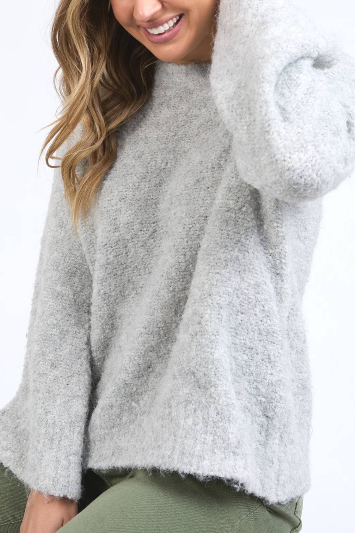 A woman smiles and touches her hair while wearing the Foxwood Mindie Knit Crew in soft, light gray with olive green pants against a plain background.