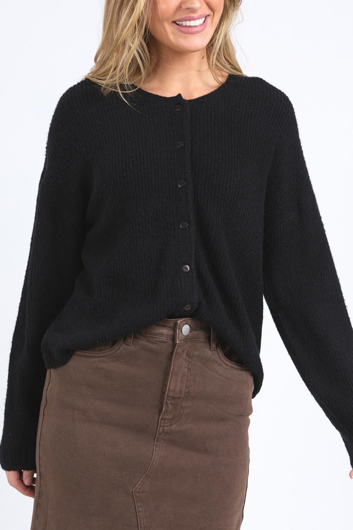 A woman smiles with her hair down, wearing the black Foxwood Murphy Knit Cardi with ribbed trims and a high-waisted brown skirt against a plain white background.