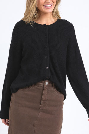 A woman smiles with her hair down, wearing the black Foxwood Murphy Knit Cardi with ribbed trims and a high-waisted brown skirt against a plain white background.