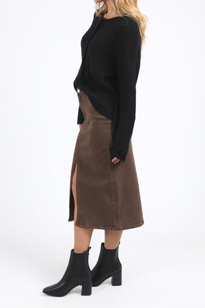 A woman stands against a white background, wearing the Foxwood Murphy Knit Cardi in black with ribbed trims, paired with a brown midi skirt featuring a front slit and black ankle boots. Her loose blonde hair obscures her face.