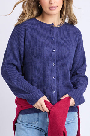 A woman wears a blue Foxwood Murphy Knit Cardi and light blue jeans, with a red sweater tied around her waist. Her hair is loose, and she stands against a plain light backdrop.