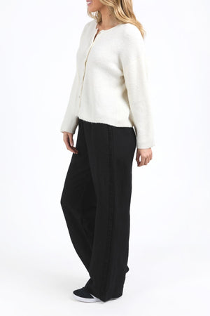 A woman stands sideways, smiling against a white background, wearing the Foxwood Murphy Knit Cardi in white, relaxed-fit black pants with a side stripe, and black slip-on shoes. She has light brown hair and looks cozy and stylish.