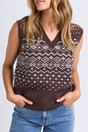 A woman with wavy light brown hair wears the Foxwood Nordic Vest, a brown knitted sleeveless sweater with a white geometric pattern, paired with blue jeans and her hands in her pockets.