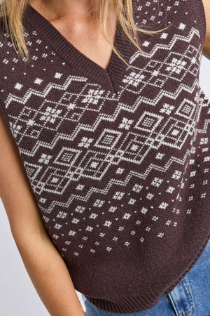 Someone wearing the Foxwood Nordic Vest, a brown knit with a white geometric and snowflake pattern, paired with blue jeans. The image is cropped to show the torso and part of one arm.