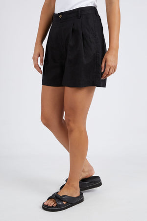Dressed in black pleated Foxwood Oakleigh Shorts featuring an elastic waistband, paired with a white top and black slip-on sandals, the person stands against a plain white background.