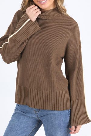 A woman wearing the Foxwood Oliver Knit, a brown ribbed sweater with wide sleeves, white arm stripe, and mock neck, pairs it with blue jeans and smiles while holding the collar near her chin for a relaxed look.