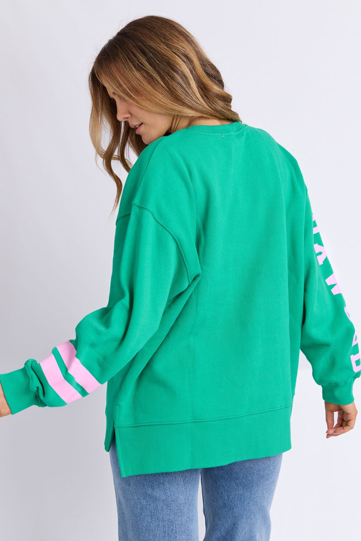 A woman with long hair faces away, wearing the Foxwood Origin Crew—a bright green relaxed-fit fleece with pink and white sleeve stripes—styled casually with light blue jeans, set against a plain background.