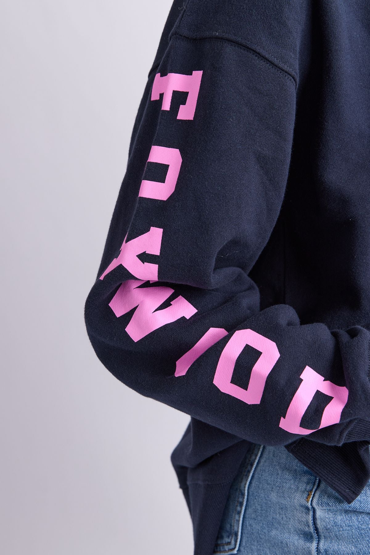 A person wears a Foxwood Origin Crew navy fleece sweatshirt with bold pink "HOLLYWOOD" lettering down the sleeve, styled with blue jeans.