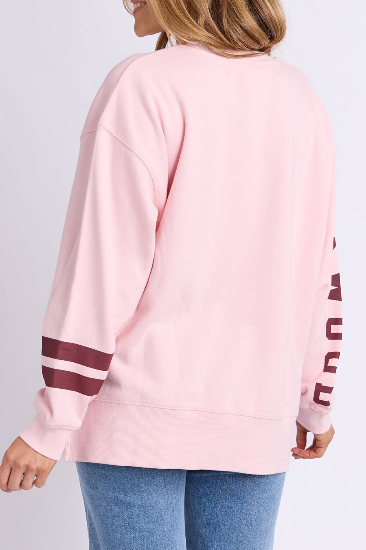 A woman seen from the back wears the Foxwood Origin Crew, a light pink relaxed-fit fleece with maroon striped sleeves and blue jeans. The sweatshirt offers casual comfort and she has loose hair.