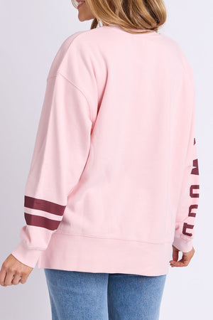 A woman seen from the back wears the Foxwood Origin Crew, a light pink relaxed-fit fleece with maroon striped sleeves and blue jeans. The sweatshirt offers casual comfort and she has loose hair.