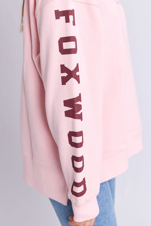Someone wears the Foxwood Origin Crew, a relaxed-fit fleece in light pink with "FOXWOOD" boldly printed vertically in maroon on the sleeve, styled with light blue jeans.
