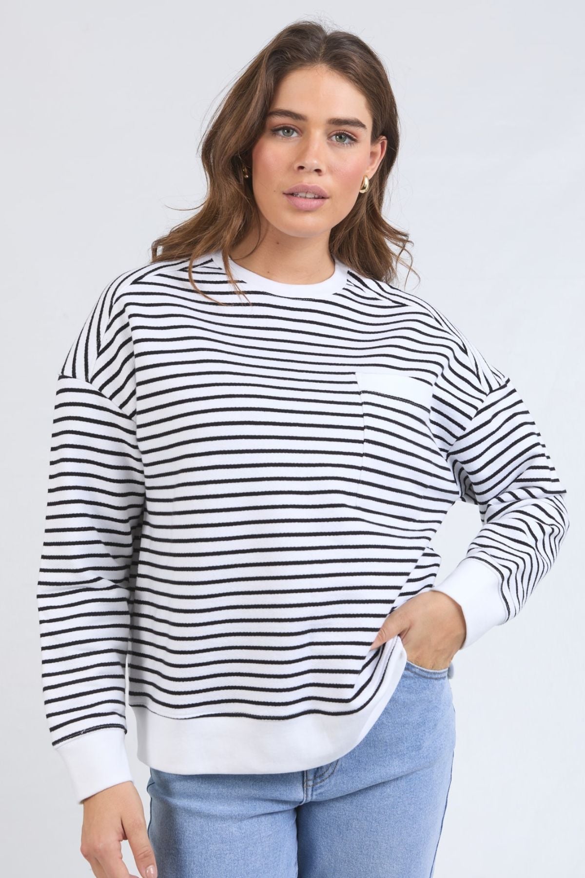 A woman in the Foxwood Original Stripe Crew, a relaxed fit jumper with cuffed sleeves, pairs it with light blue jeans and stands with one hand on her hip, facing the camera against a plain light background.