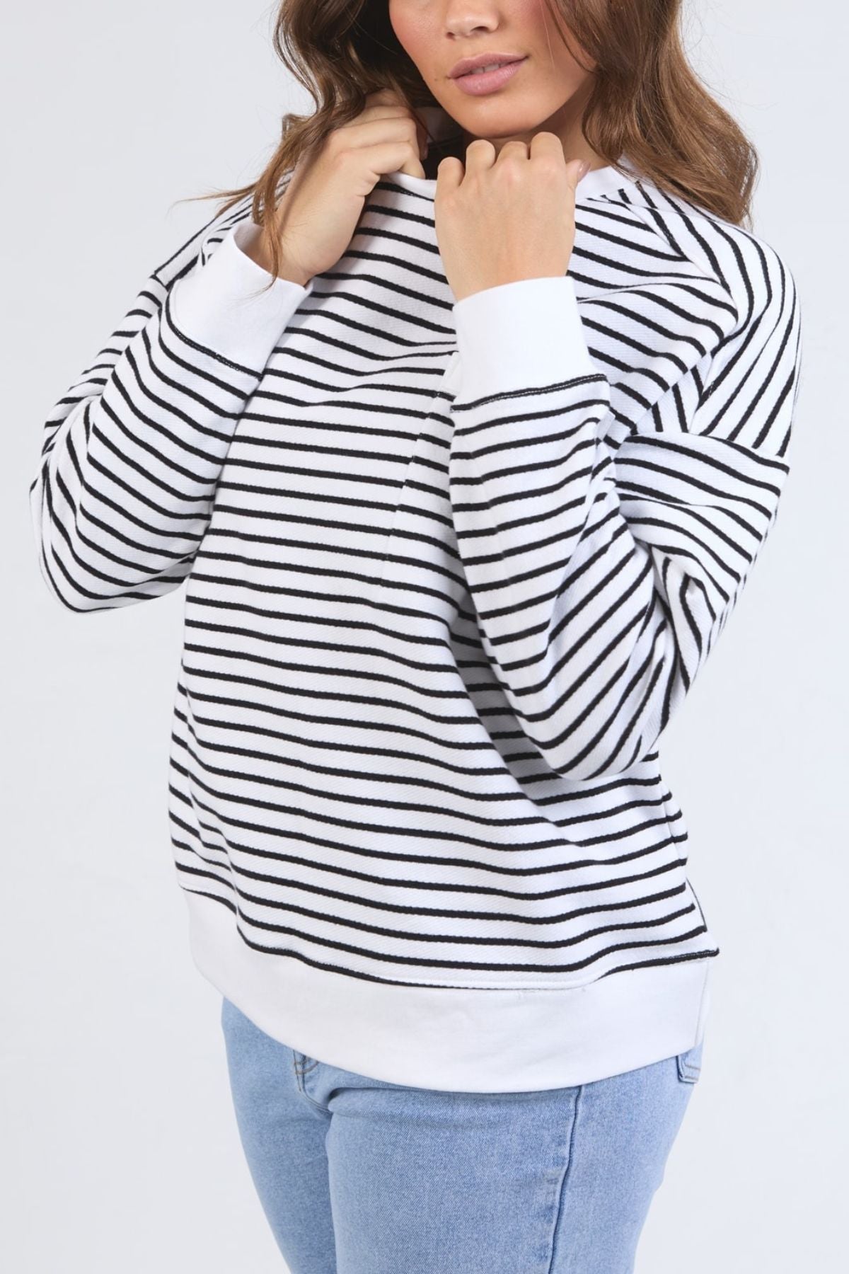 Wearing the Foxwood Original Stripe Crew relaxed fit jumper with cuffed sleeves, paired with light blue jeans, a woman stands against a plain light background, lightly pulling at the collar as her face is partially visible.