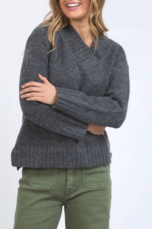 A woman with wavy blonde hair smiles in the Foxwood Pepper Knit, a cozy sweater with long sleeves and relaxed fit, paired with olive green pants. She stands with arms crossed against a light background.