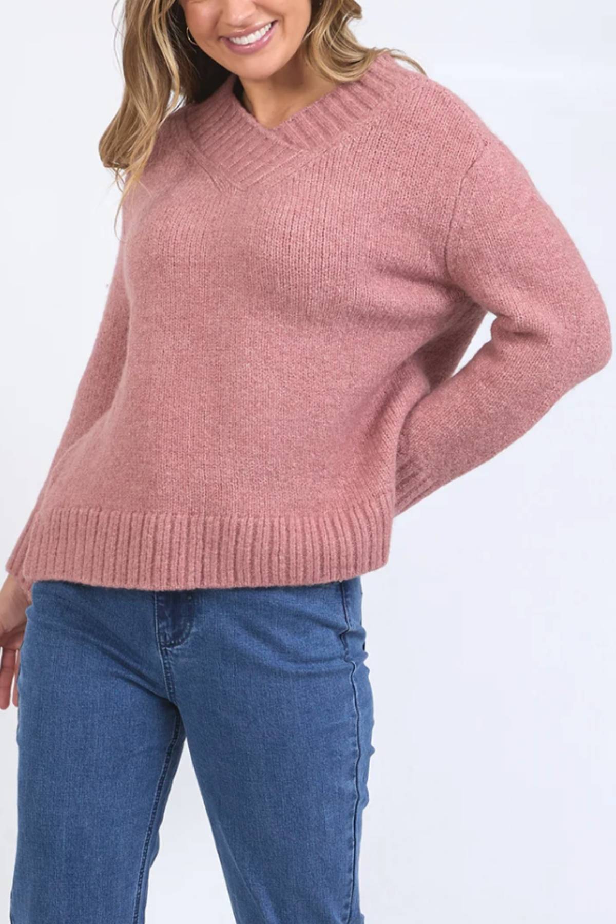 A person wearing a relaxed fit, dusty pink Foxwood Pepper Knit V-neck sweater and blue jeans stands with one hand on their hip, smiling.