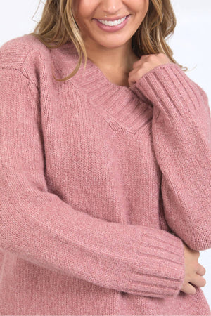 A person smiles while wearing the Foxwood Pepper Knit, a cozy pink long-sleeved sweater in a relaxed fit, with one hand near their face and loose wavy hair partly visible.