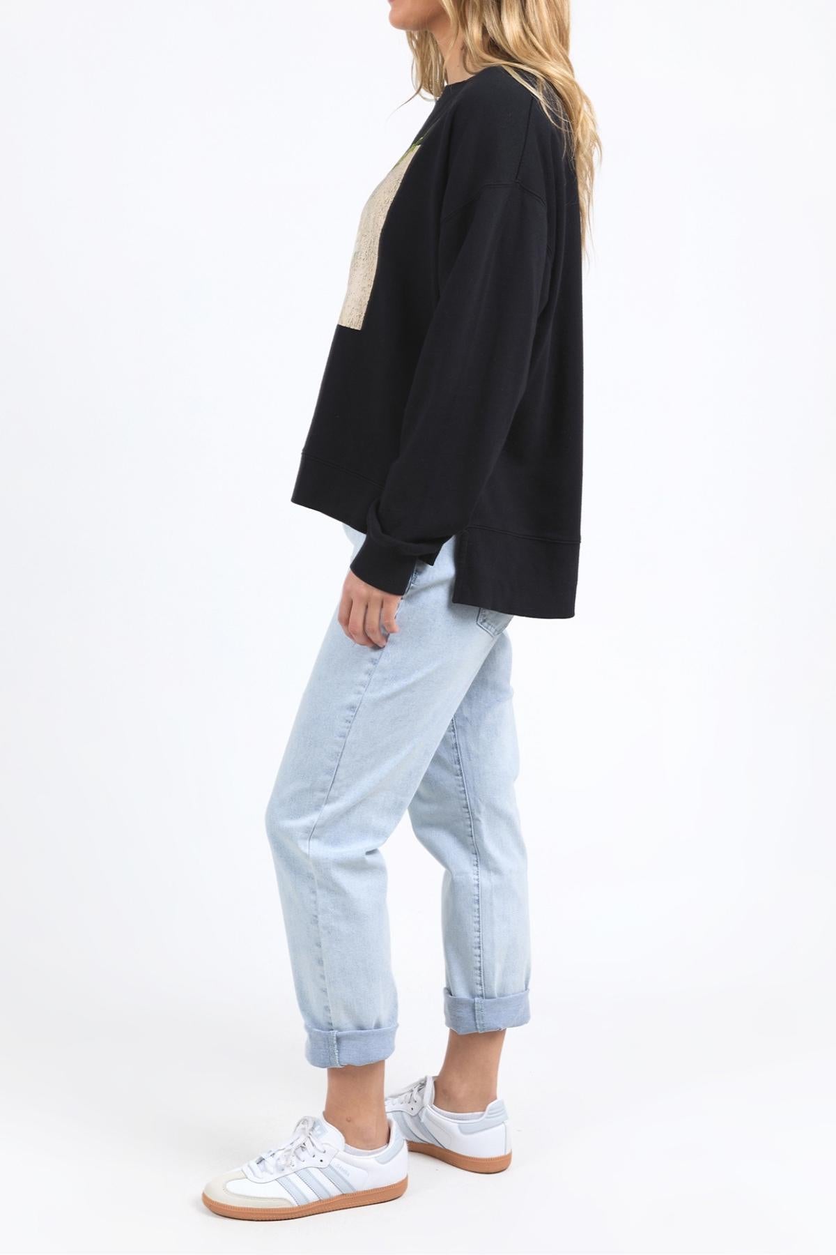 A woman stands sideways against a white background, wearing the Foxwood Posy Crew with light blue cuffed jeans and white sneakers. For sizing, check the half chest measurement or contact our customer service team for help.