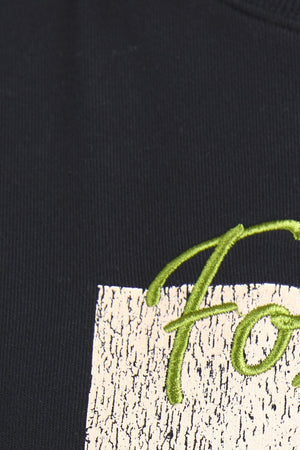 Close-up of the black Foxwood Posy Crew fabric featuring a cream-colored, textured rectangle with part of “Forest” embroidered in green. For sizing help, our customer service team is ready to assist you.