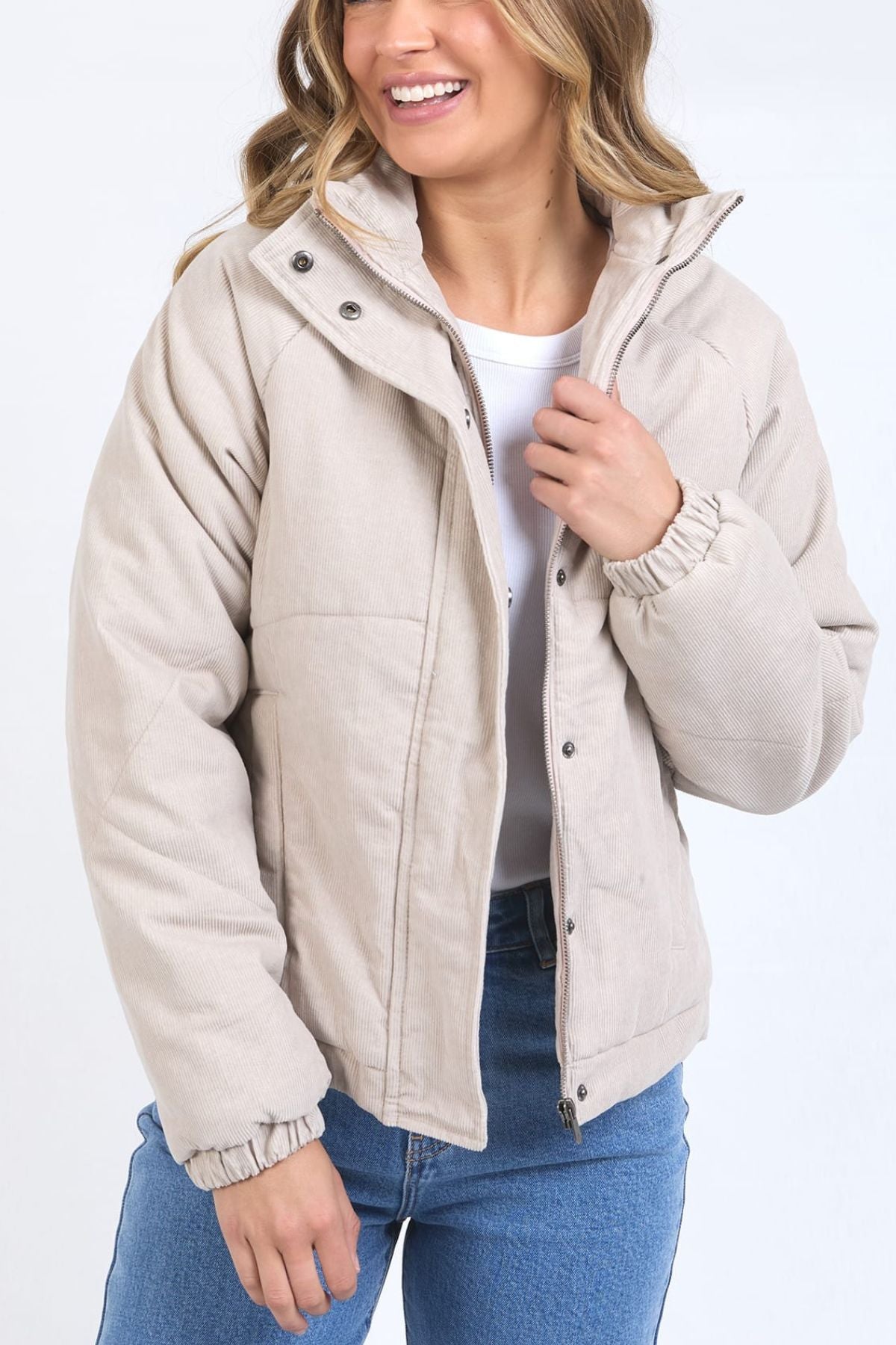 A woman wearing the Foxwood Rosalee Cord Jacket in light beige over a white shirt and blue jeans, smiling and holding the jacket collar with one hand against a plain white background.