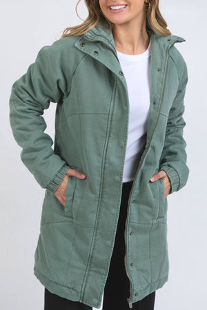 A smiling woman wears the Foxwood Rosalee Longline Jacket in light green with snap buttons over a white shirt, hands in jacket pockets, standing against a plain light background for a cozy yet stylish look.
