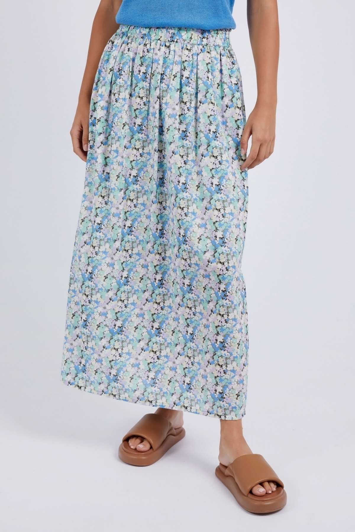 A person wears a blue top and the Foxwood Rosette Floral Skirt, featuring a luxe cotton poplin with light blue, green, and white floral pattern, paired with tan slip-on sandals against a plain white background.