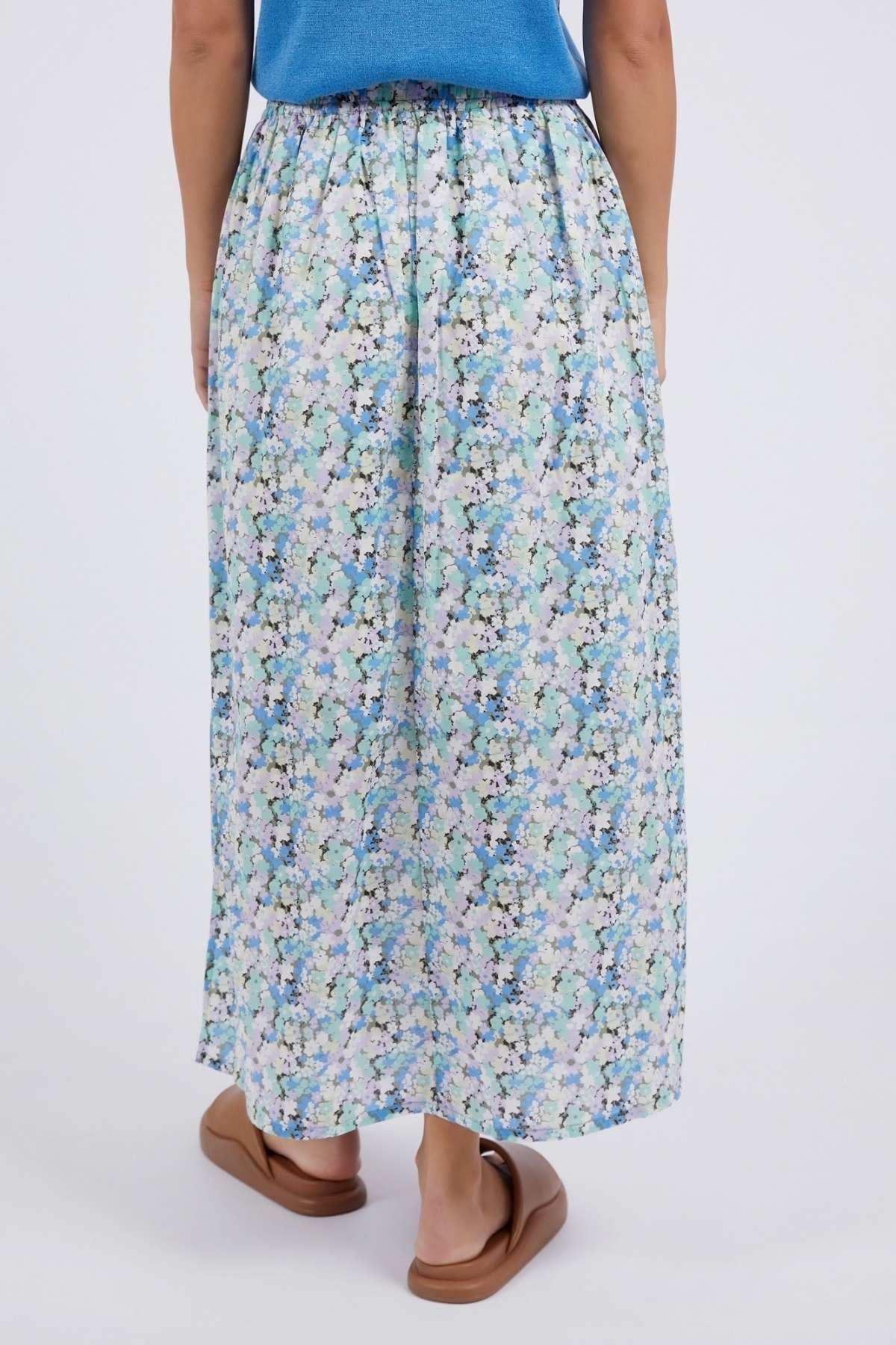 A person is shown from the back wearing a blue top, brown sandals, and the Foxwood Rosette Floral Skirt with a vibrant blue, green, and white print against a plain light background.