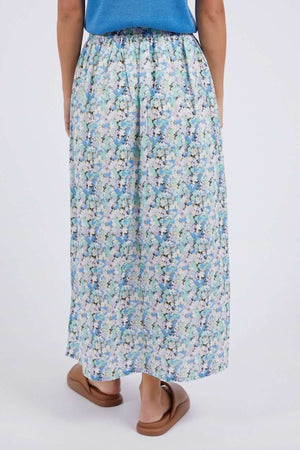 A person is shown from the back wearing a blue top, brown sandals, and the Foxwood Rosette Floral Skirt with a vibrant blue, green, and white print against a plain light background.