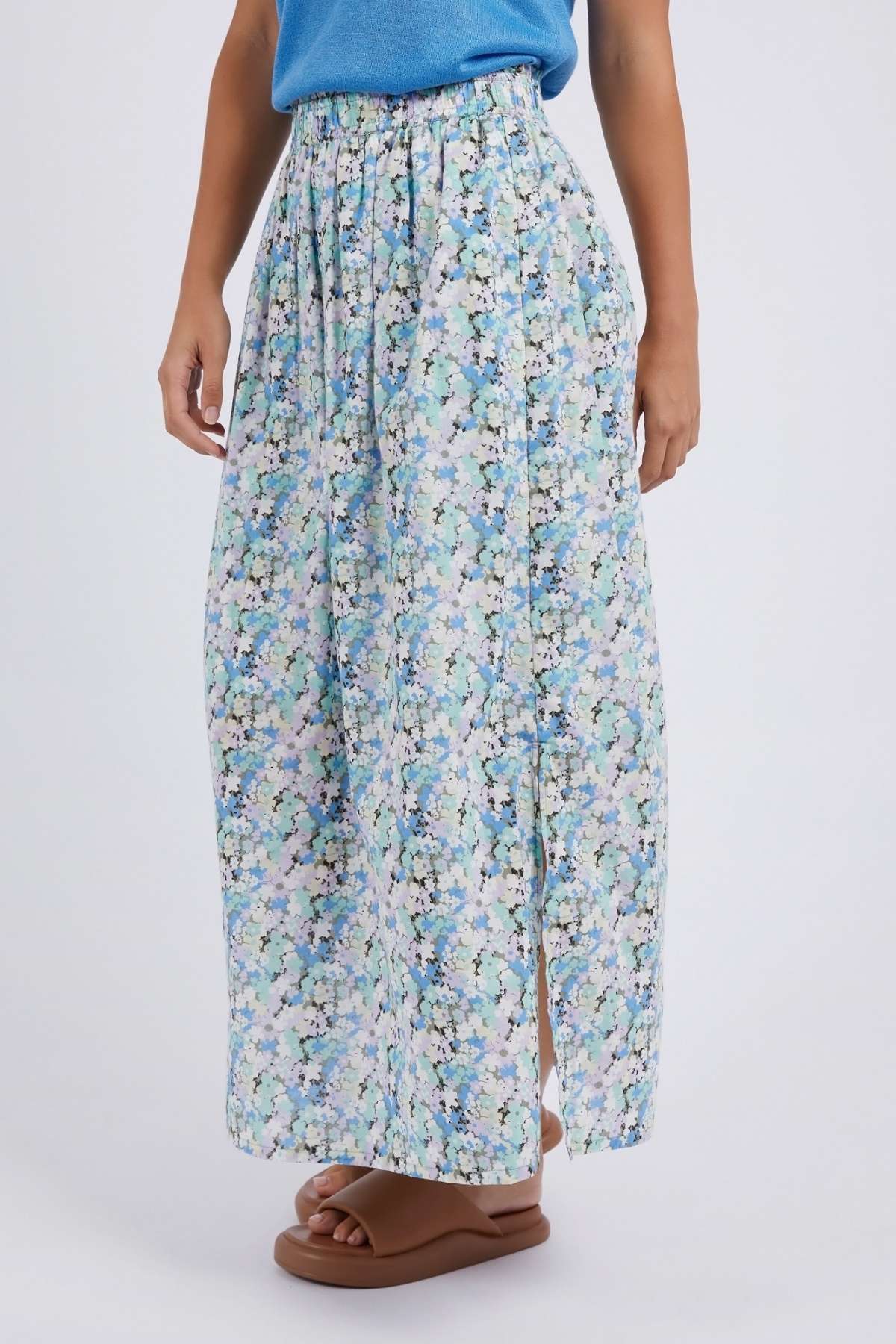 Someone wears the Foxwood Rosette Floral Skirt, a long, high-waisted floral skirt with a side slit and vibrant print, styled with a blue top and brown sandals against a plain white background.