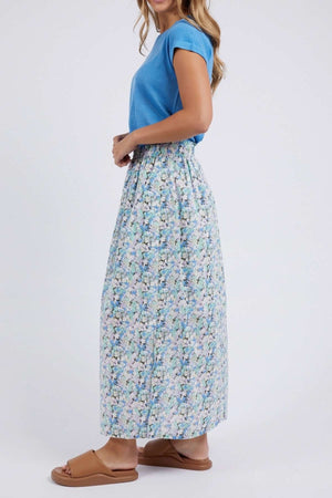 A woman stands sideways in a Luxe Cotton Poplin blue short-sleeve top, Foxwood Rosette Floral Skirt with vibrant light blue, green, and white florals, and tan flat sandals. The background is plain white.