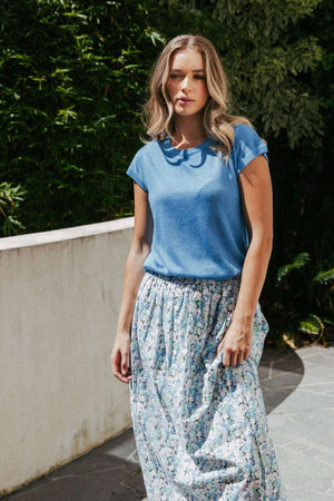 A woman with wavy blonde hair wears a blue short-sleeve top and the Foxwood Rosette Floral Skirt, a long, flowy piece in luxe cotton poplin, standing outdoors near a white wall with greenery in the background.