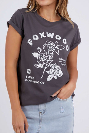 A woman models the Foxwood Rosette Tee, a relaxed fit dark gray 100% cotton top with white rose graphics and “FOXWOOD” on the front, styled with a light blue denim skirt.