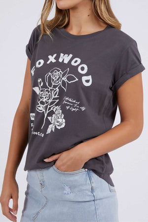 A woman wears the Foxwood Rosette Tee, a dark gray cotton T-shirt featuring a white rose graphic and relaxed fit, paired with a light blue denim skirt. She stands with one arm relaxed; her face is partially out of frame.