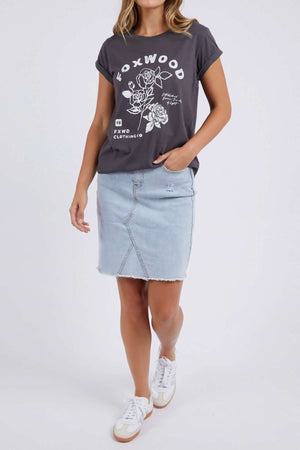 A woman models the Foxwood Rosette Tee, a dark graphic t-shirt featuring white floral designs and lettering, styled with a light blue denim skirt and white sneakers against a plain backdrop.