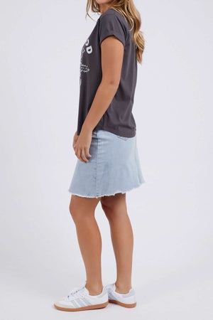 A woman stands sideways wearing the Foxwood Rosette Tee, a black relaxed-fit graphic shirt, paired with a light blue frayed-hem denim skirt and white gum-soled sneakers against a plain white background.