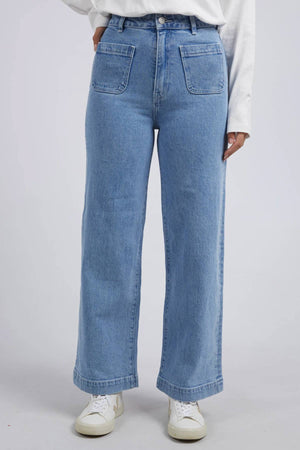 A person wears the Foxwood Sarah Wide Leg Jean, a high-rise, light blue wide leg denim with large front pockets, paired with a white long-sleeve shirt and white sneakers, standing against a plain light background.