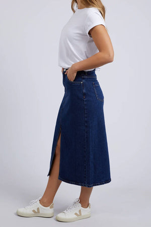 A woman stands in profile against a plain light background, wearing a white t-shirt, white sneakers, and the Foxwood Scout Midi Skirt in light blue stretch denim with a front slit.