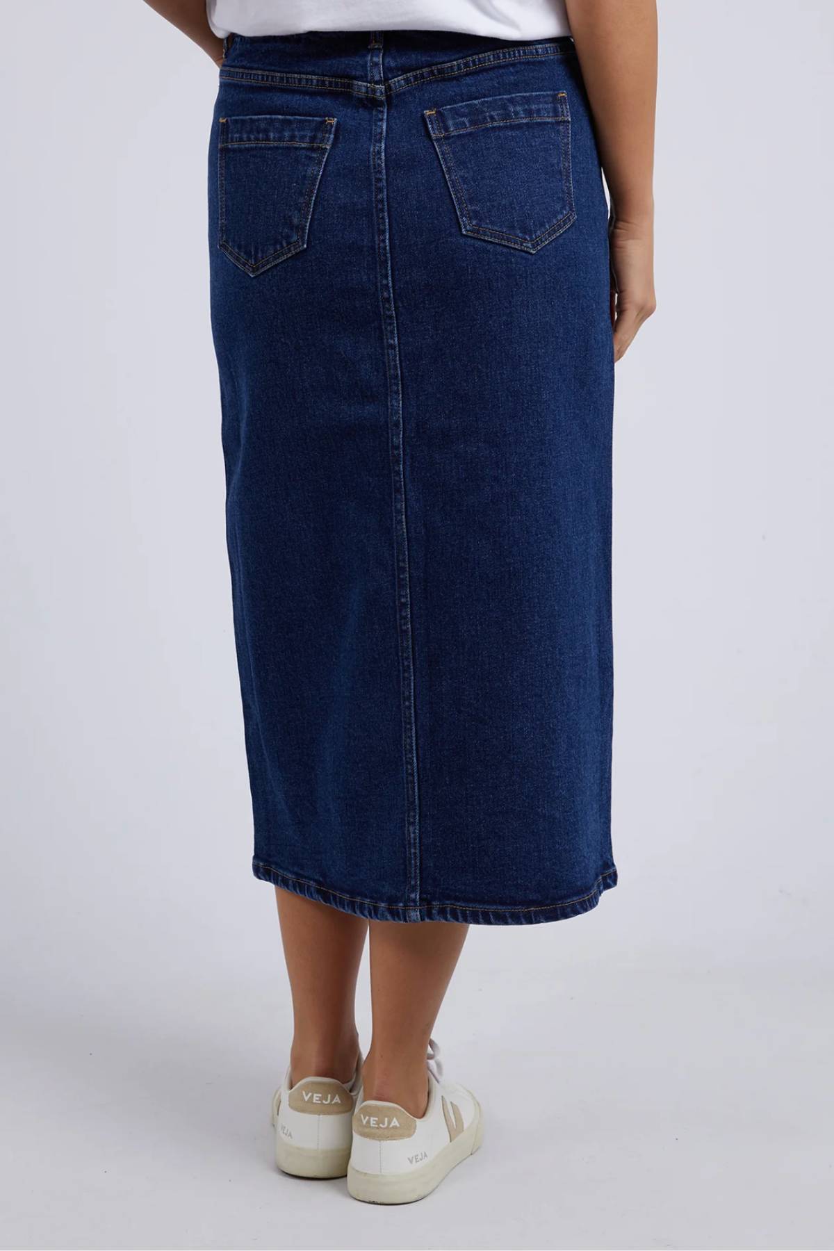 A person wearing a light blue Foxwood Scout Midi Skirt and white Veja sneakers stands with their back to the camera against a plain light background.