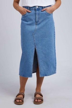 A person models the chic Foxwood Scout Midi Skirt in blue denim featuring a front slit, paired with a white top and brown sandals. Standing stylishly against a neutral background, they casually place their hands in the skirt's pockets.