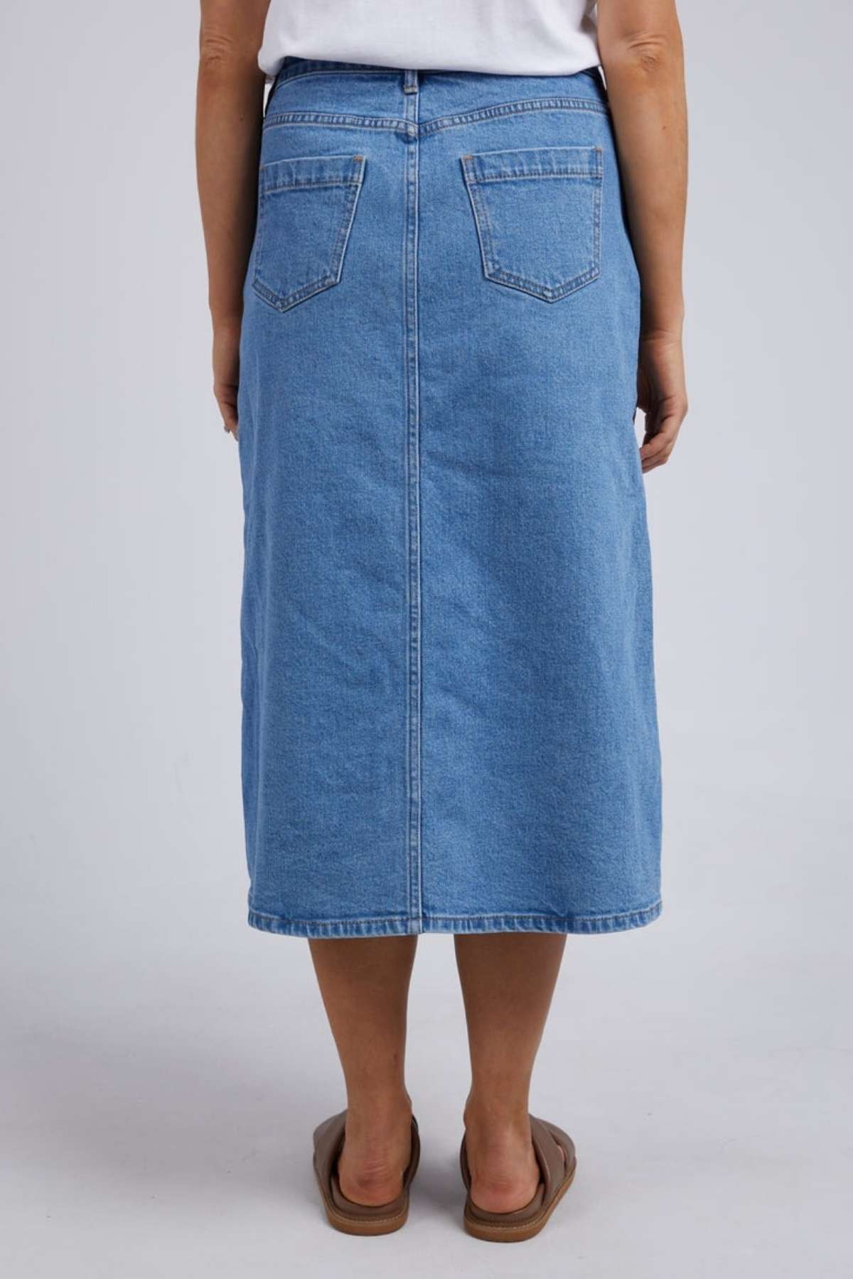 A person is seen from behind wearing the Foxwood Scout Midi Skirt in blue vintage denim, paired with a white top and brown sandals against a plain backdrop—a stylish ensemble capturing timeless fashion.