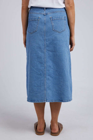 A person is seen from behind wearing the Foxwood Scout Midi Skirt in blue vintage denim, paired with a white top and brown sandals against a plain backdrop—a stylish ensemble capturing timeless fashion.