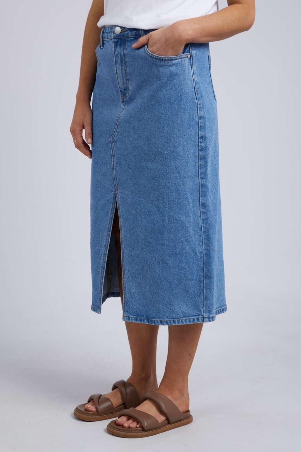 A trendy person wears a white top with the Foxwood Scout Midi Skirt, high-waisted and knee-length with a front slit, paired with brown sandals on a light gray background, achieving a chic vintage denim style.
