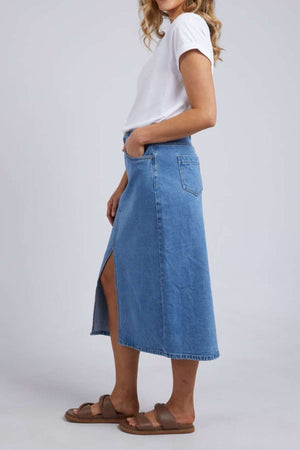 Wearing a white t-shirt and the stylish Foxwood Scout Midi Skirt, noted for its side slit, someone stands sideways with hands in pockets. The outfit is paired with brown sandals, all set against a simple light background.