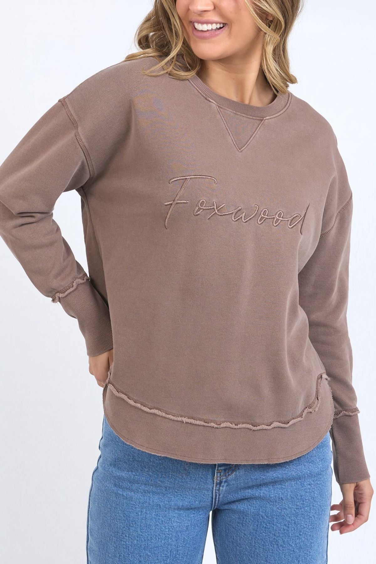A woman wears the Foxwood Signature Crew, a 100% cotton crewneck with "Foxwood" embroidered on the front, paired with blue jeans. She smiles confidently and poses with one hand on her hip.