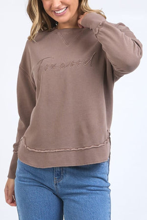 A woman in the Foxwood Signature Crew—a 100% cotton crewneck sweater with "Foxwood" embroidery—pairs it with blue jeans, smiling as she touches her hair against a plain light background.