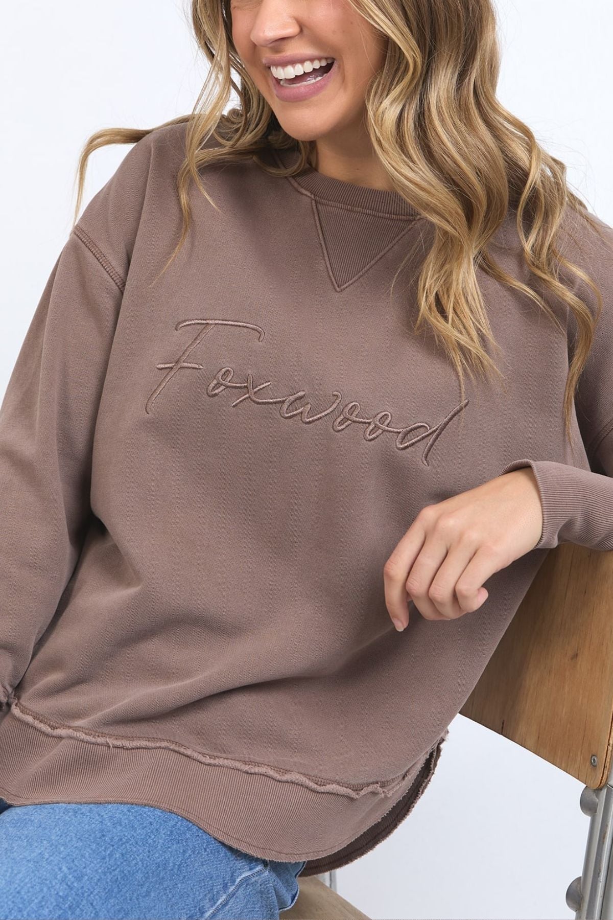 A woman with wavy blonde hair sits smiling on a wooden chair, wearing the Foxwood Signature Crew—a brown 100% cotton crewneck sweater featuring "Foxwood" embroidery—paired with light blue jeans.