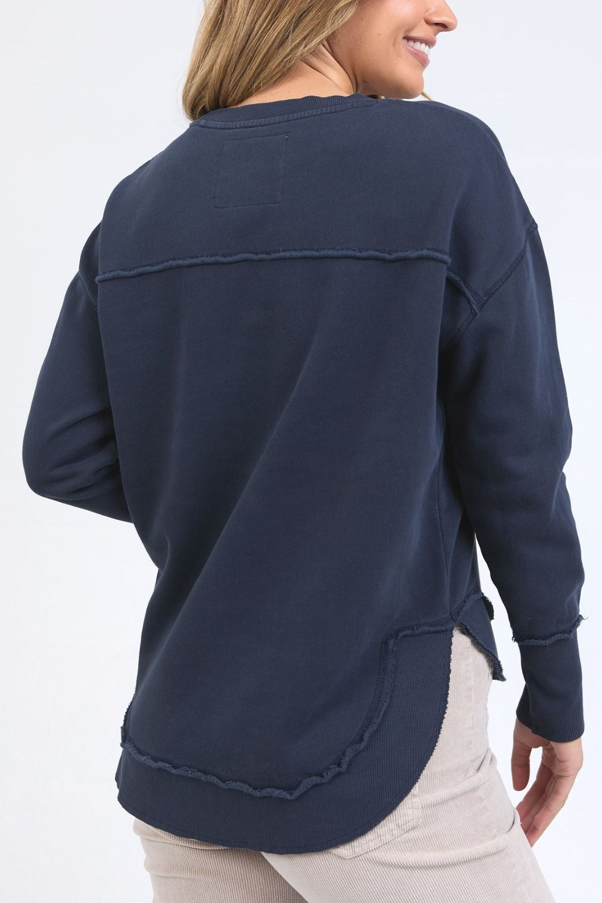 A woman with light brown hair and light-colored pants smiles as she stands with her back to the camera, wearing the Foxwood Signature Crew—a navy blue, 100% cotton long-sleeve crewneck sweater with raw-edge seams and a curved hem.
