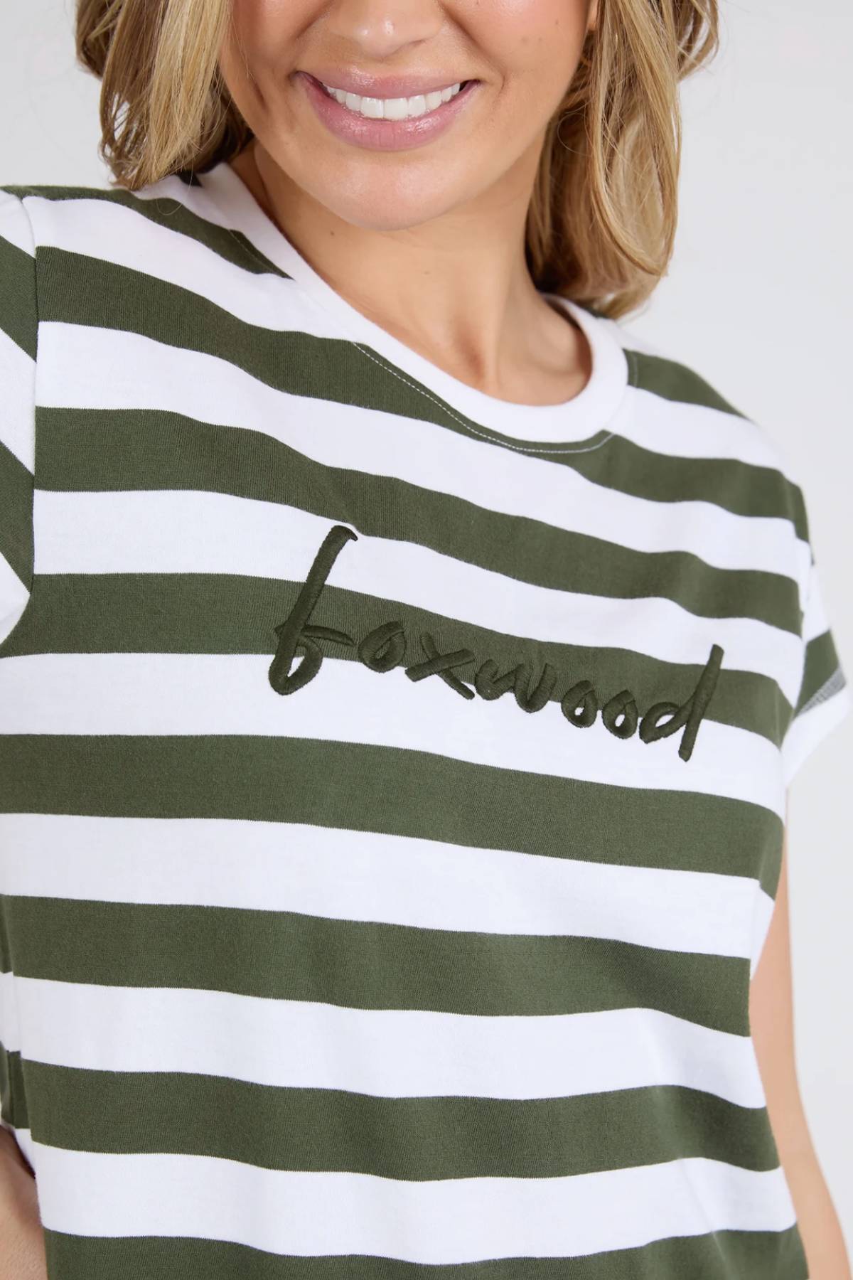 Woman smiling and wearing the Foxwood Signature Stripe Tee Dress—a white and olive green striped t-shirt dress with the Foxwood logo embroidered in black script across the chest.