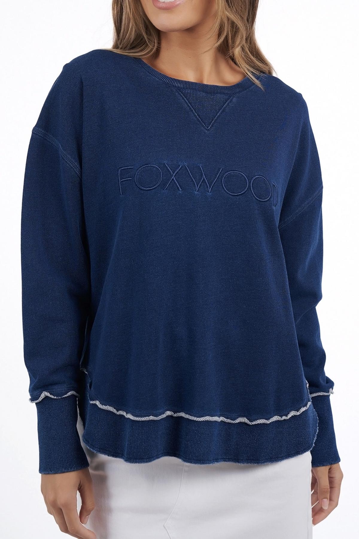 A woman models the Foxwood Simplified Crew, a navy 100% cotton sweatshirt with "FOXWOOD" embroidery and white trim, paired with a white skirt. Her face is partially out of frame.