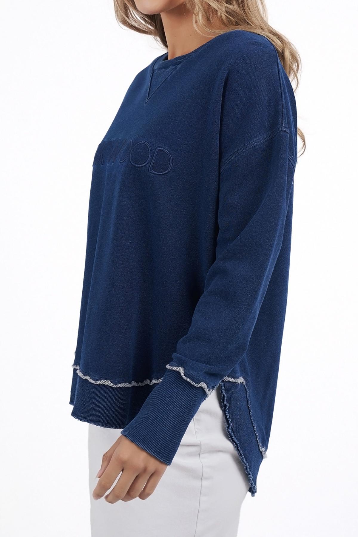 Someone wearing a relaxed fit, long-sleeve Foxwood Simplified Crew in navy blue with subtle chest text and frayed edges stands against a white background, paired with white pants.